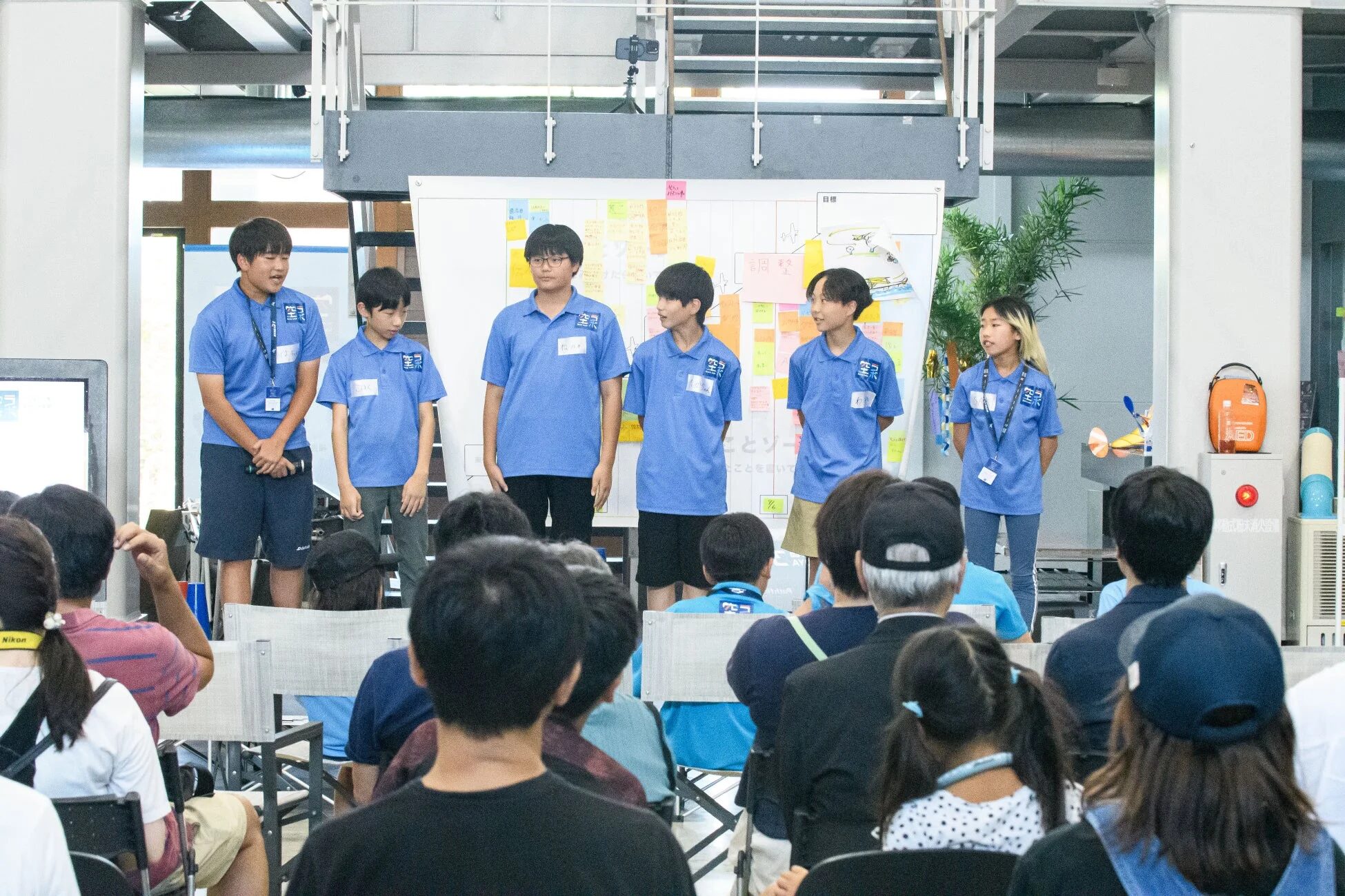 青いポロシャツを着た学生たちが、付箋が貼られたホワイトボードの前で発表している様子