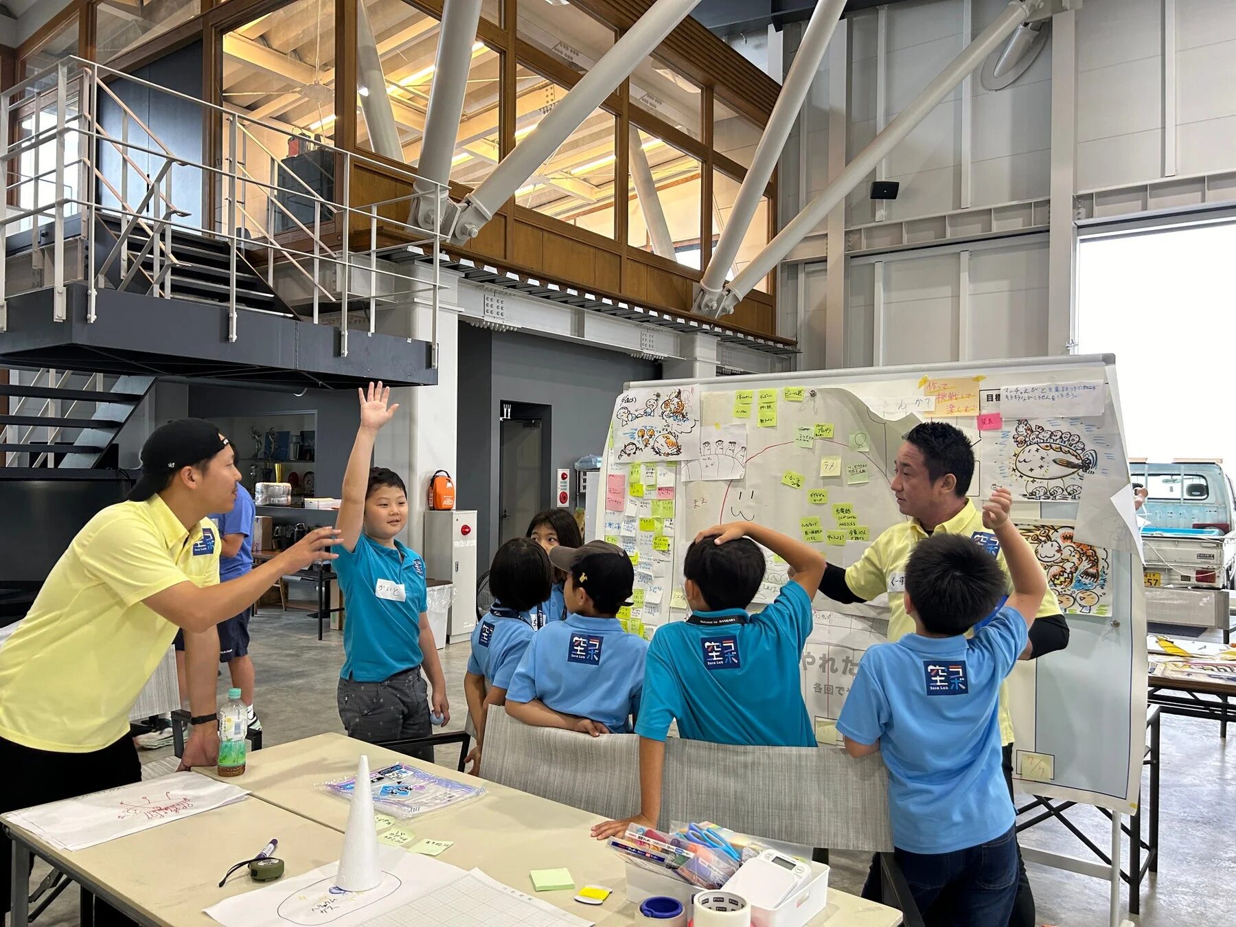 青いシャツを着た子供たちが、黄色いシャツの大人からホワイトボードを使って説明を受けているワークショップの様子
