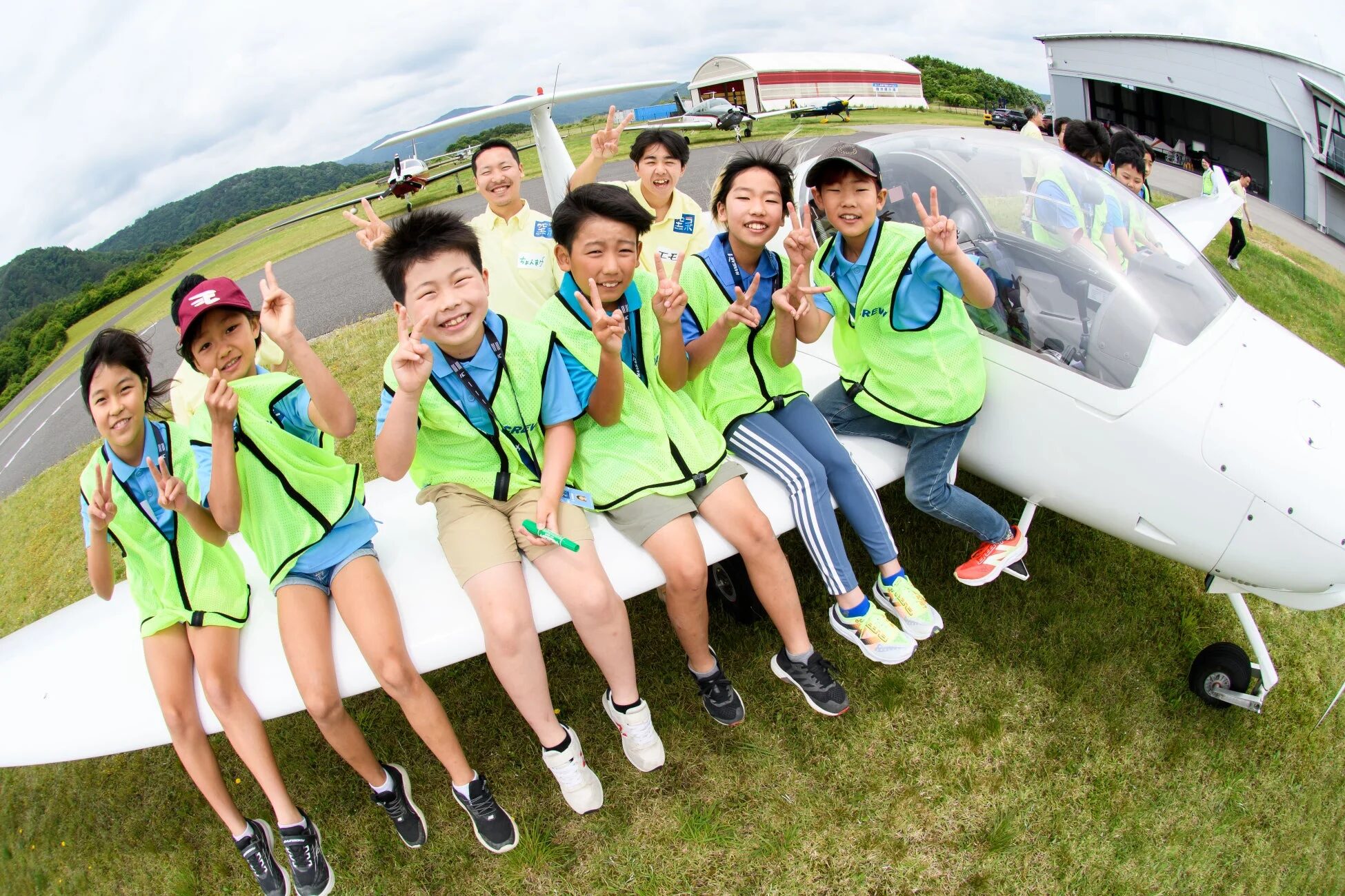 飛行場で白いグライダーの翼に座り、ピースサインをして笑顔を見せる子供たちと数人の大人たちのグループ写真