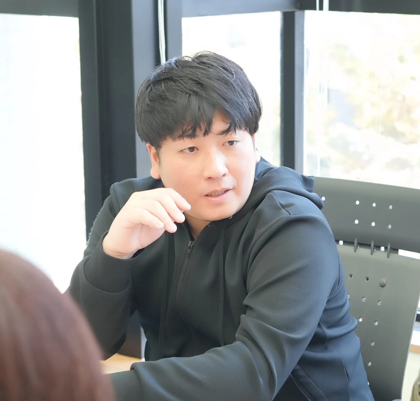黒いフード付きジャケットを着た若い男性が、室内で椅子に座り、真剣な表情で話を聞いている様子