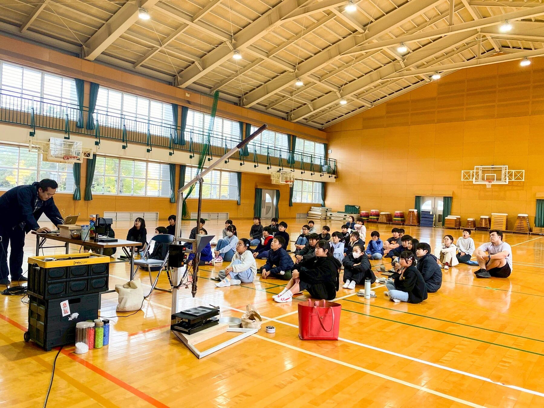 体育館で行われているワークショップの様子。講師が機材を使い実演する中、床に座った生徒たちが熱心に見学しています。