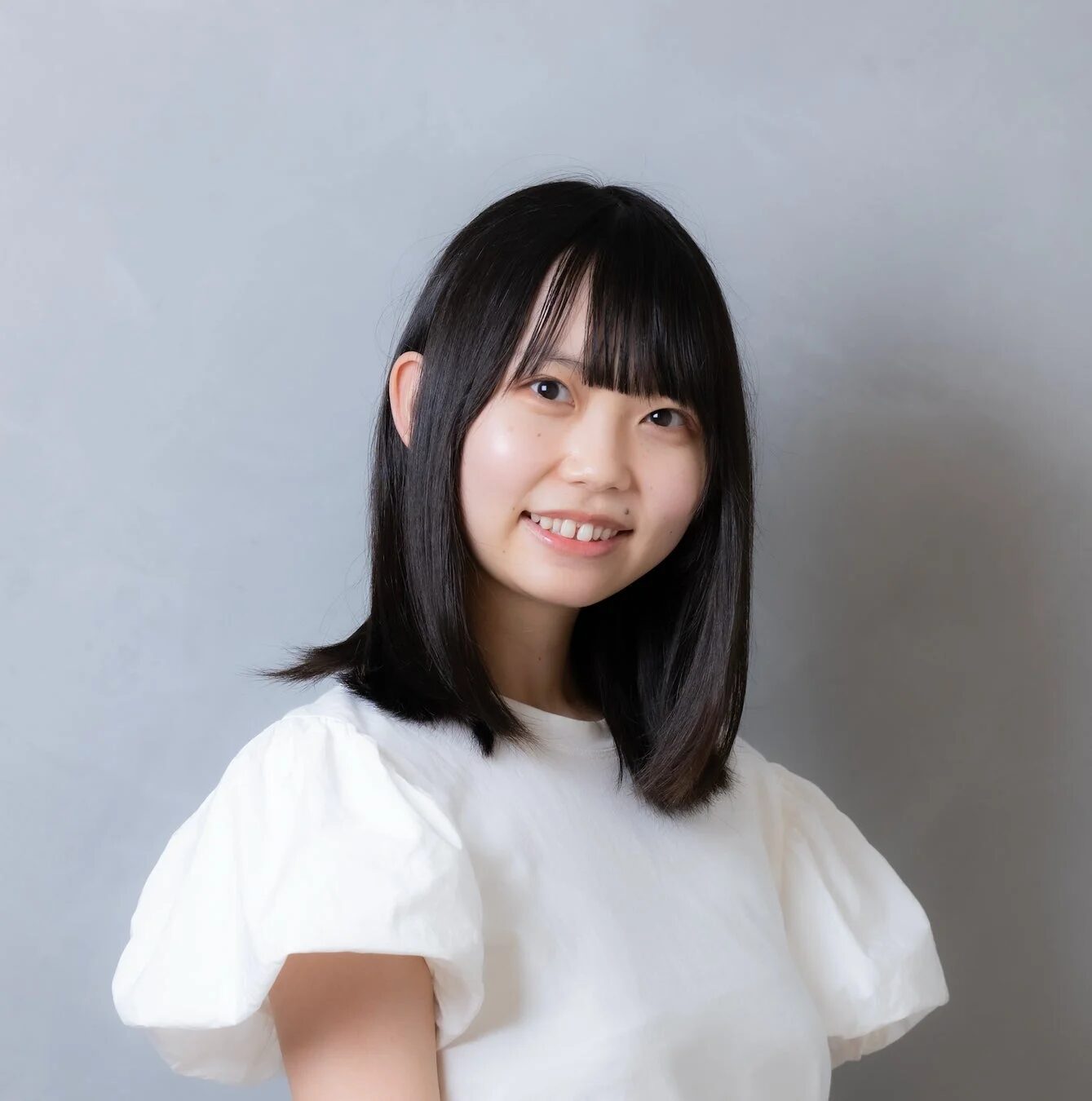 グレーの背景の前で、白いパフスリーブのトップスを着た若いアジア人女性が微笑んでいます