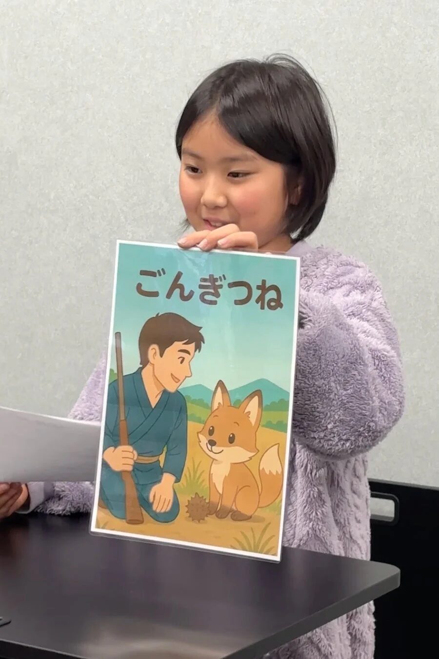 ふわふわの紫色の服を着た若い女の子が、「ごんぎつね」という文字と、男性とキツネが描かれたイラストボードを笑顔で持っています。何かの発表をしている場面のようです。