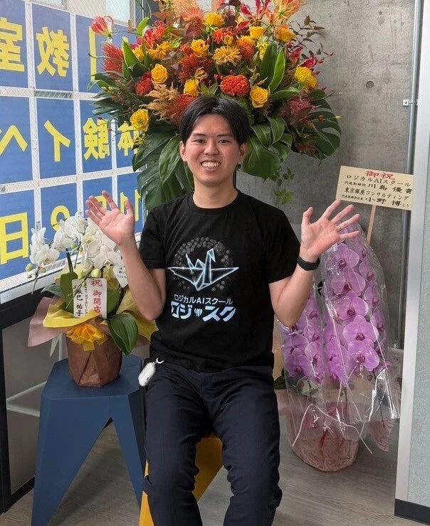 ロジカルAIスクールのTシャツを着た男性が、開店祝いの豪華な花と胡蝶蘭の前で笑顔で両手を広げています。壁の文字や祝札から、新しいAIスクールの開校を祝う場面であることがわかります。