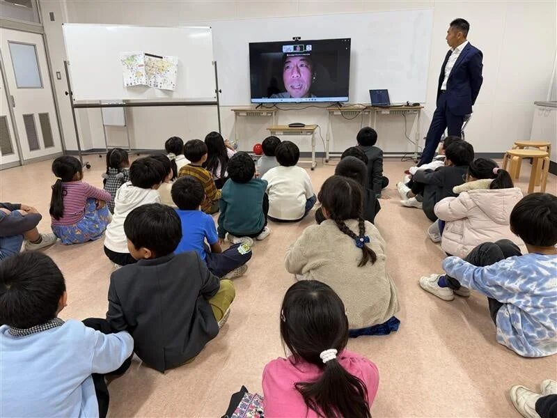 大型スクリーンに映し出された人物のオンライン講義を聴く子供たち