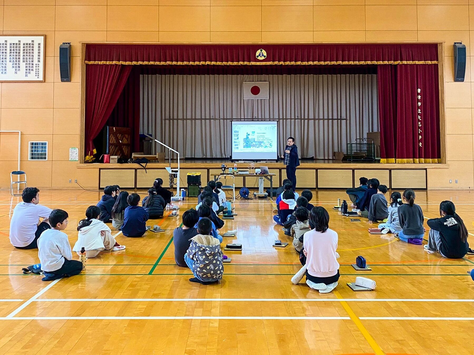 体育館で小学生たちが床に座ってステージ上の大人によるプレゼンテーションを聴いている様子。