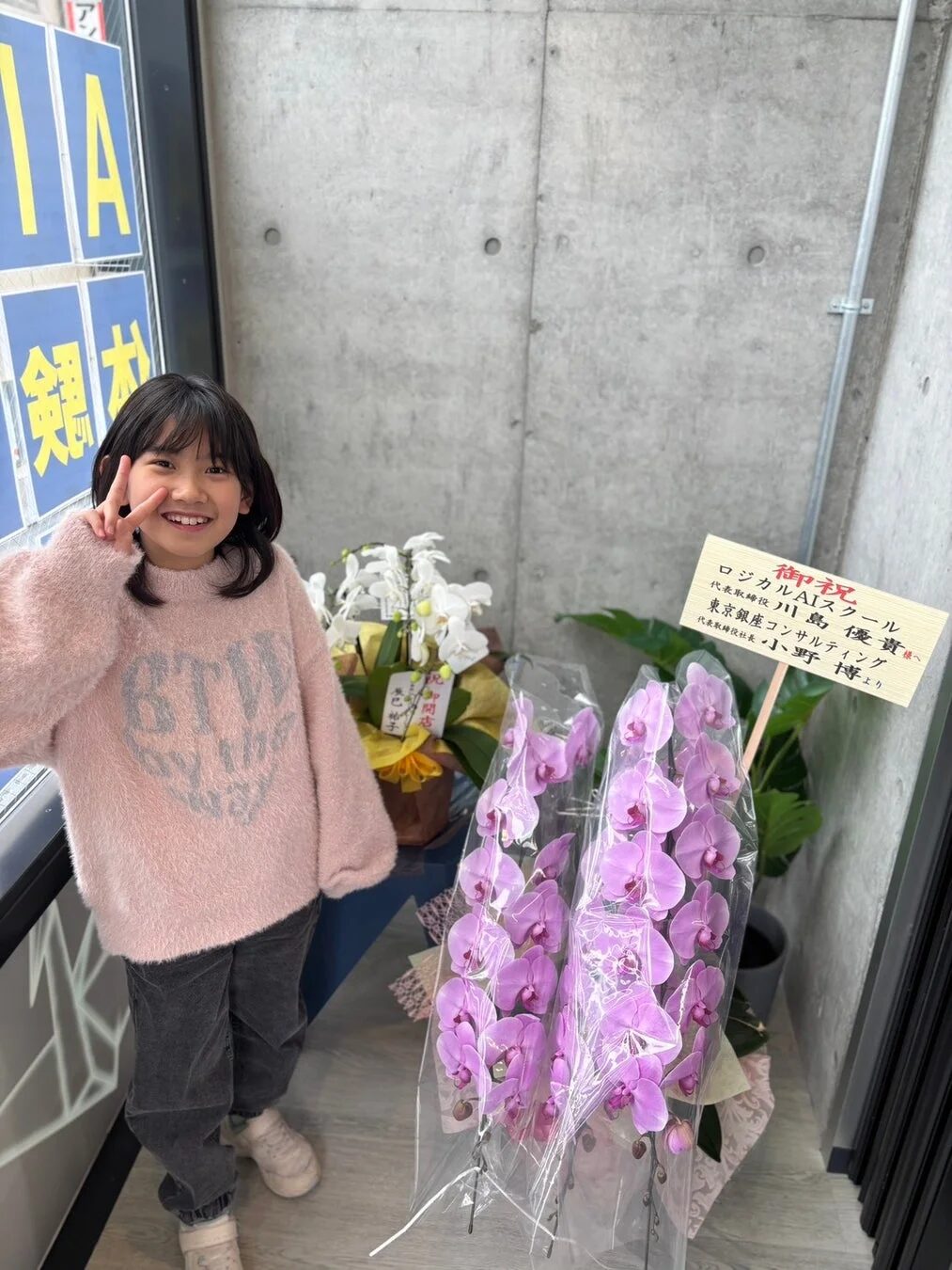笑顔でピースサインをする可愛らしい女の子が写っています。背景には「銀座」の文字と、胡蝶蘭を含むたくさんのお祝いの花が飾られており、「ロジカルAIスクール」や「東京銀座コンサルティング」への開店祝いのようです。モダンなコンクリートの壁が特徴的な室内で撮影されています。