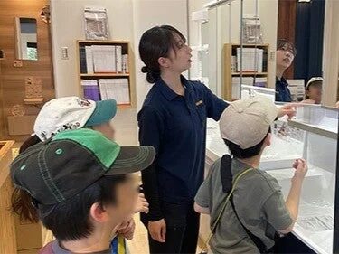 店員らしき女性が数人の子供たちに何かを説明している場面です。