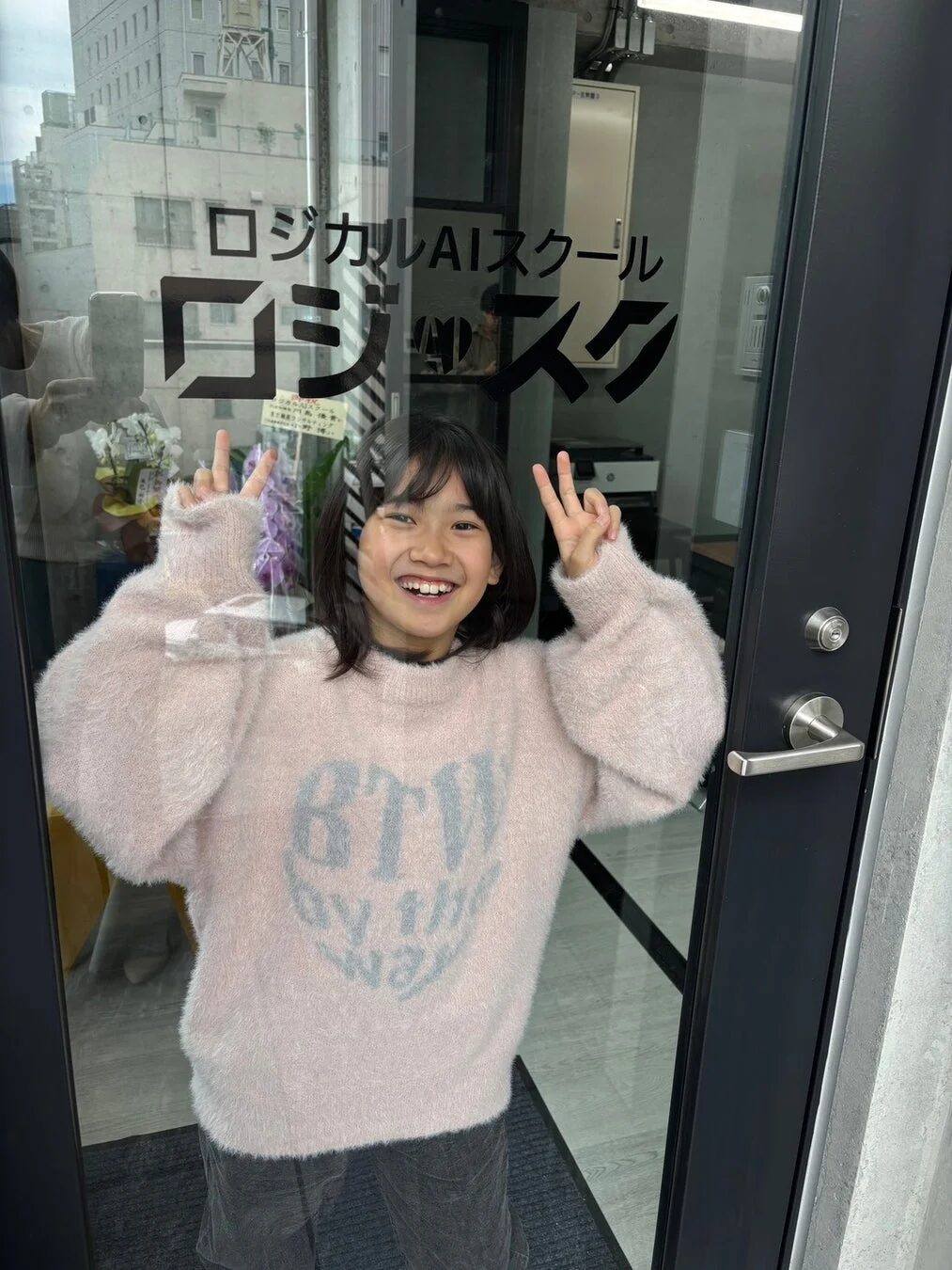 ロジカルAIスクールのガラス扉の前で、ピンクのふわふわしたセーターを着た女の子が、満面の笑みで両手でピースサインをしています。