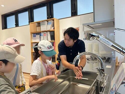 ショールームのような場所で、男性が子供たちにキッチンのシンクと蛇口の使い方を説明しています。