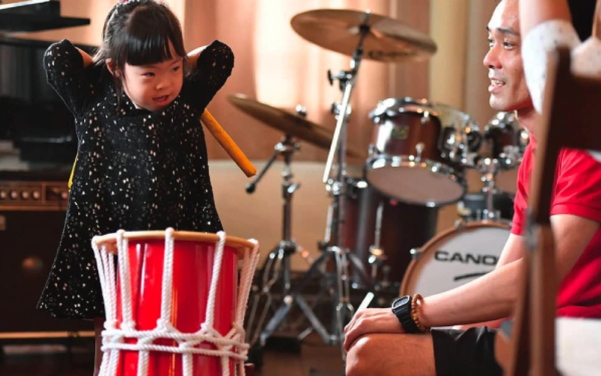 ドラムキットのあるスタジオで、小さな子供が赤い太鼓を叩き、隣で大人が優しく見守っている