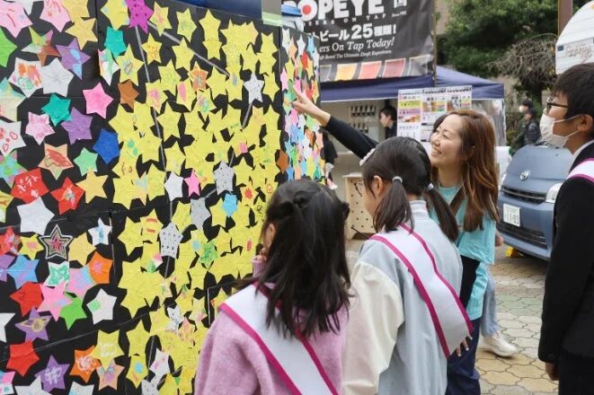 屋外イベントで、人々がカラフルな星形の紙が貼られた壁の飾り付けを眺めています