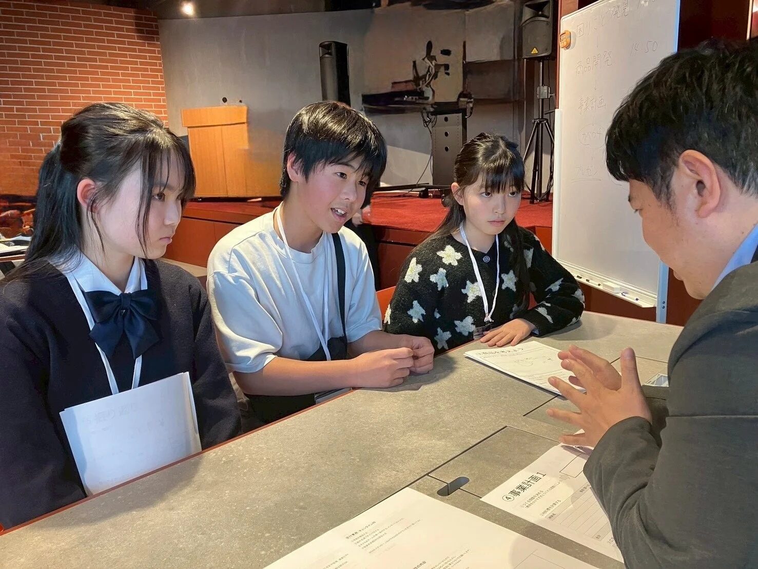 子供たちが大人から説明を受けている様子、ホワイトボードに「商品開発」「事業計画」