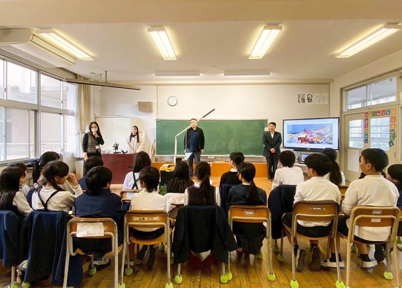 日本の小学校の教室で、児童たちが大人たちによる環境問題に関する授業を受けている様子です。スクリーンにはゴミが散乱する海岸の写真が映し出されています。