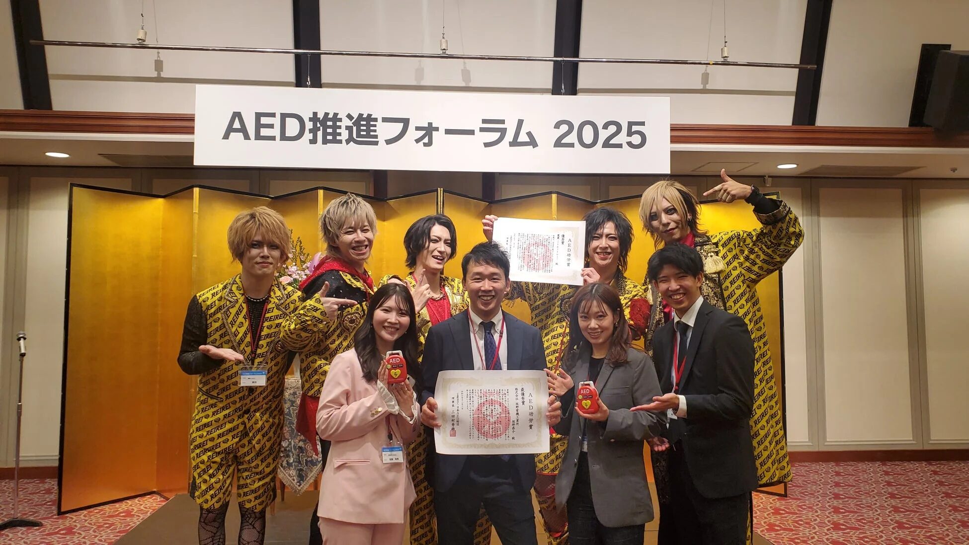 AED推進フォーラム2025の会場で、派手な衣装の男性たちとスーツ姿の男女が賞状やAEDの模型を持ち、笑顔で記念撮影をしている様子。