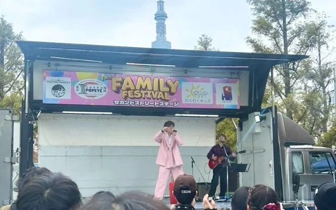 屋外のイベント会場で「FAMILY FESTIVAL」のステージが開催され、ピンクの衣装を着たシンガーとギタリストが観客の前でパフォーマンスを披露している