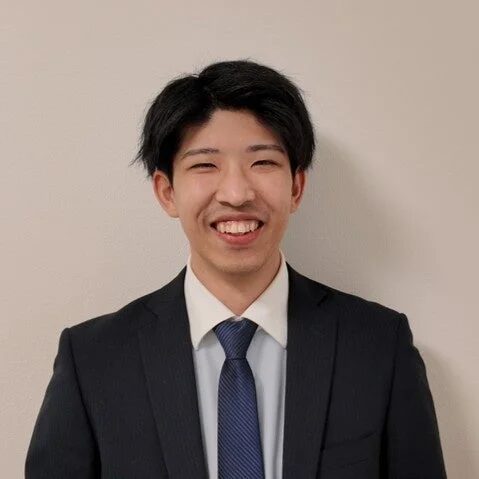 白い背景の前で、黒いスーツと青いネクタイを着用し、満面の笑みを浮かべている若いアジア人男性の上半身ポートレートです。