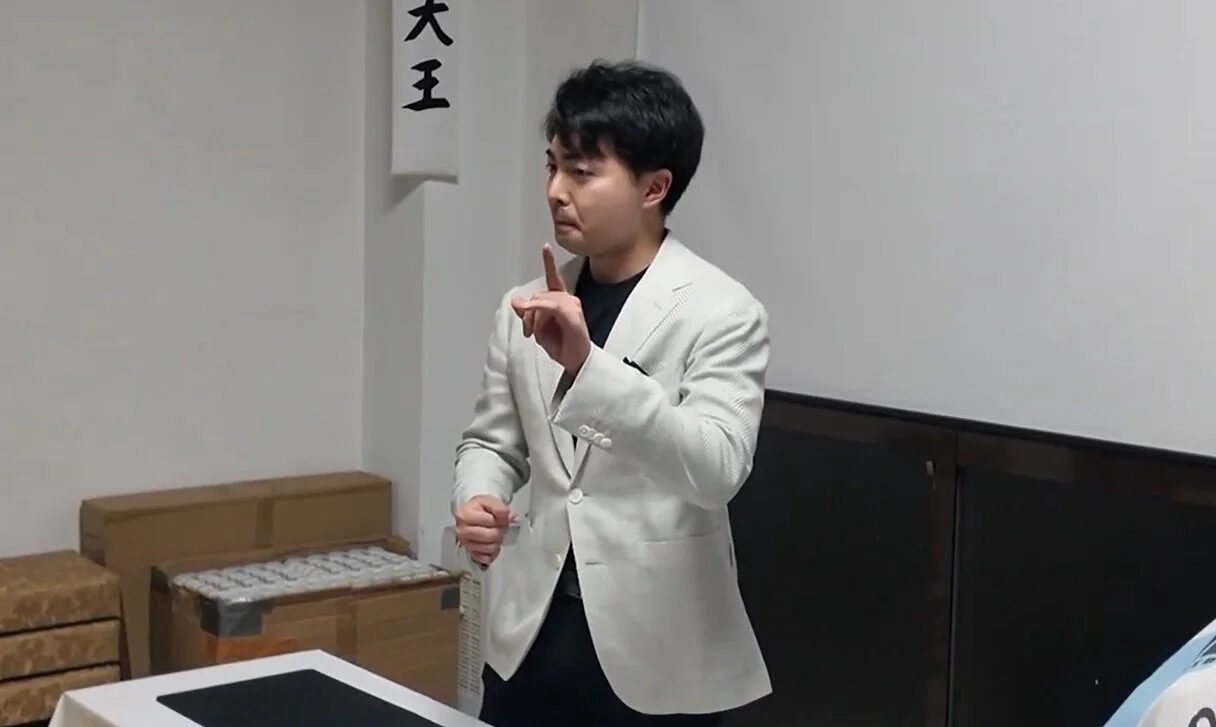 白いブレザーを着た男性が室内で指を立てて話している