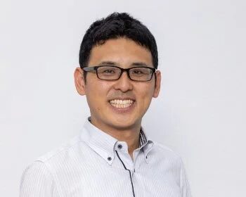 眼鏡をかけた男性が白いシャツを着て笑顔を見せているポートレート写真です。