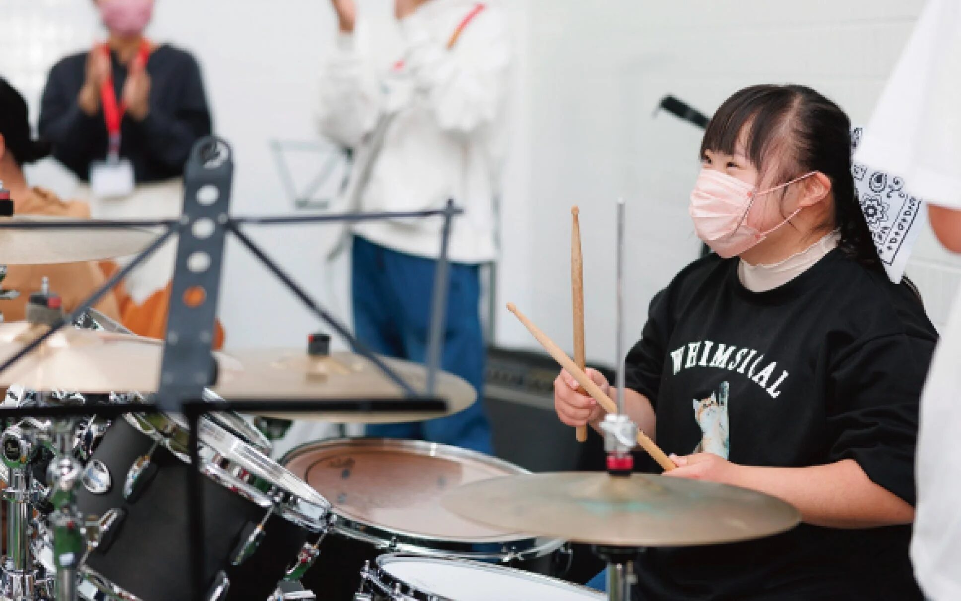 ピンクのマスクをつけた若い女性が、笑顔でドラムを演奏している