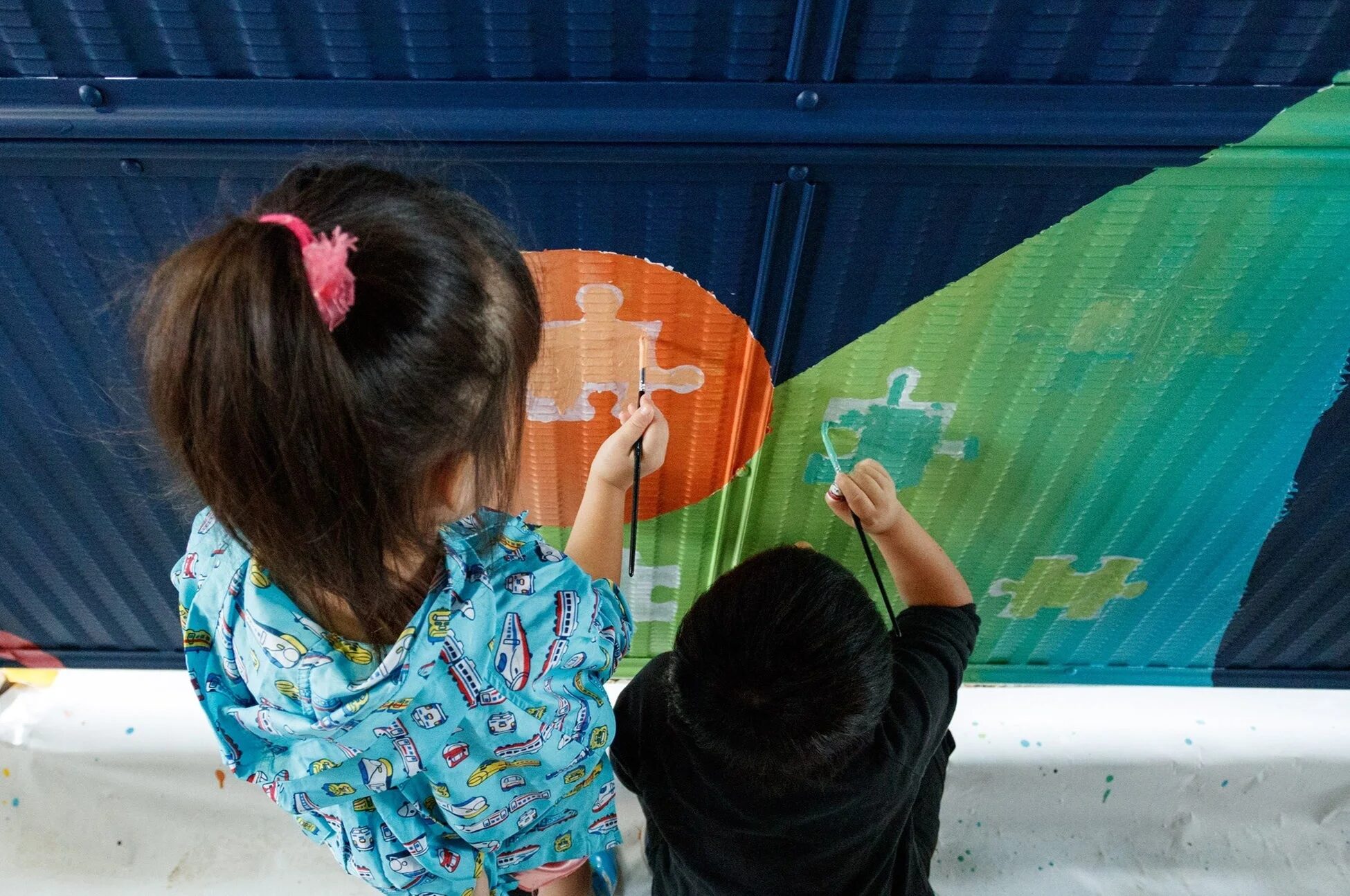 上から見た構図で、2人の子供がカラフルな波状の壁にパズルのピースの絵を描いている様子