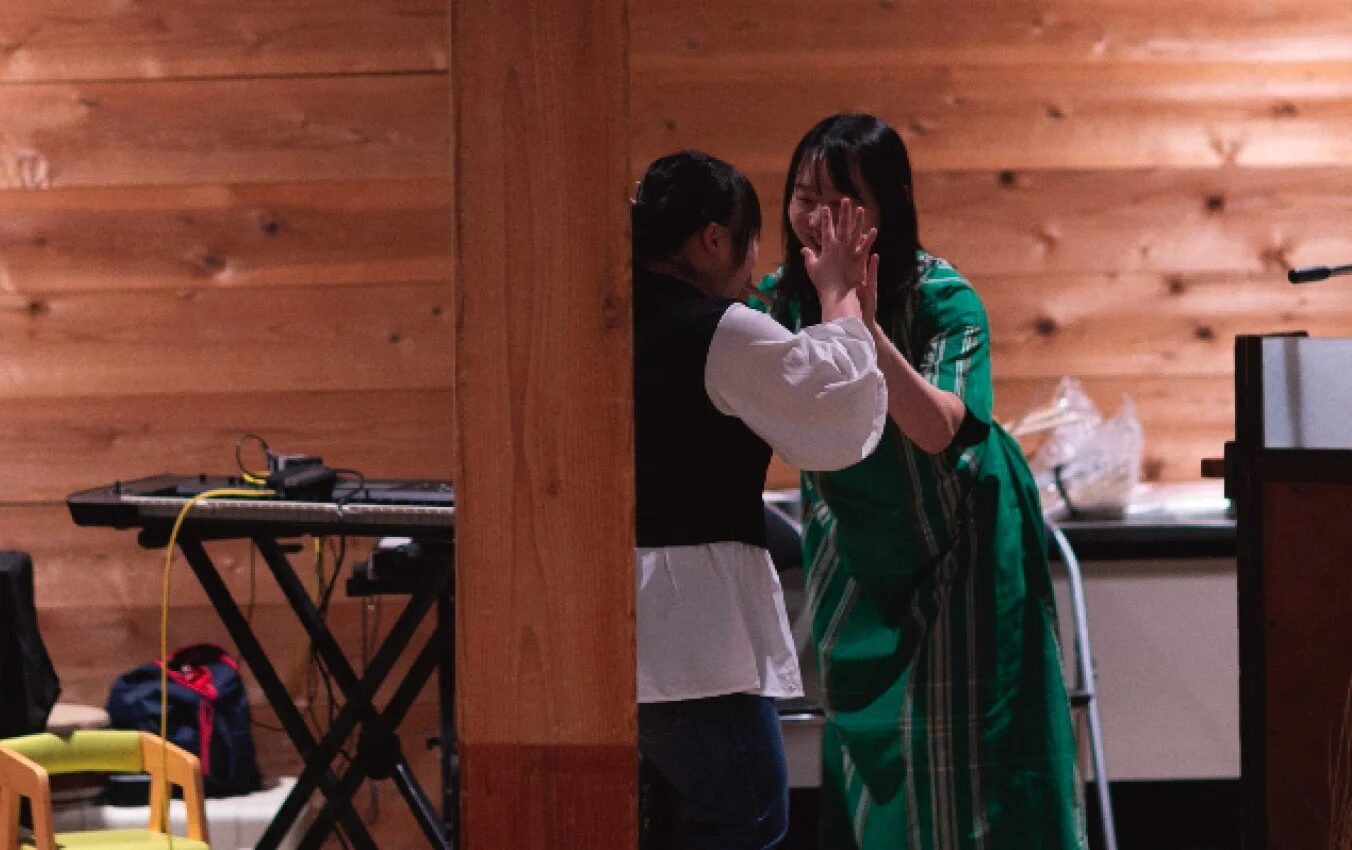 木製の壁の部屋で、二人の女性が向かい合って手を合わせ、楽しそうに笑い合っている