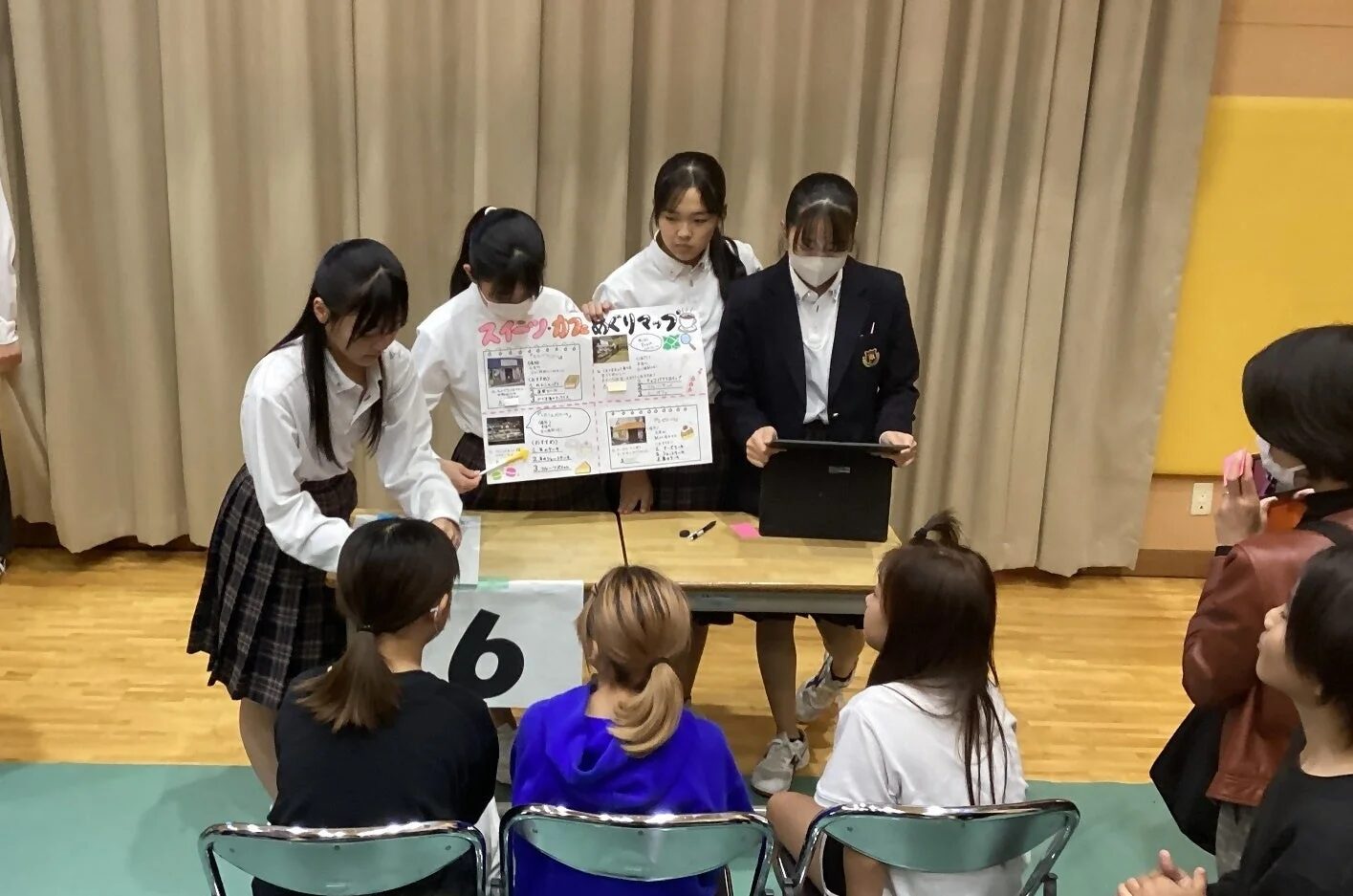 複数の女子生徒が「スイーツ・カフェめぐりマップ」と書かれたポスターを使ってプレゼンテーションを行っています。他の生徒たちが聴衆として座っており、学校の発表会やイベントの一場面と見られます。