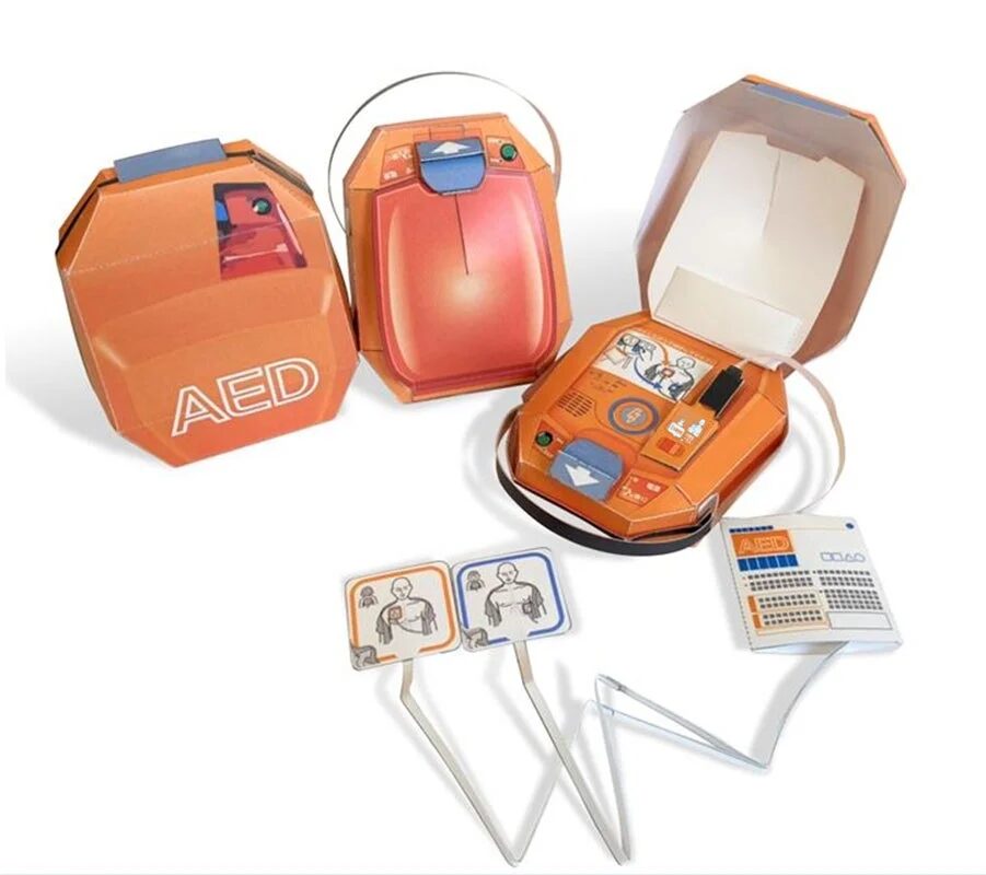 自動体外式除細動器（AED）のペーパークラフト模型が複数写っています。閉じた状態、一部開いた状態、完全に開いて内部構造や操作方法がわかる状態のAEDがあり、パッド装着位置を示す図解カードも含まれています。訓練やデモンストレーション用の教材と思われます。