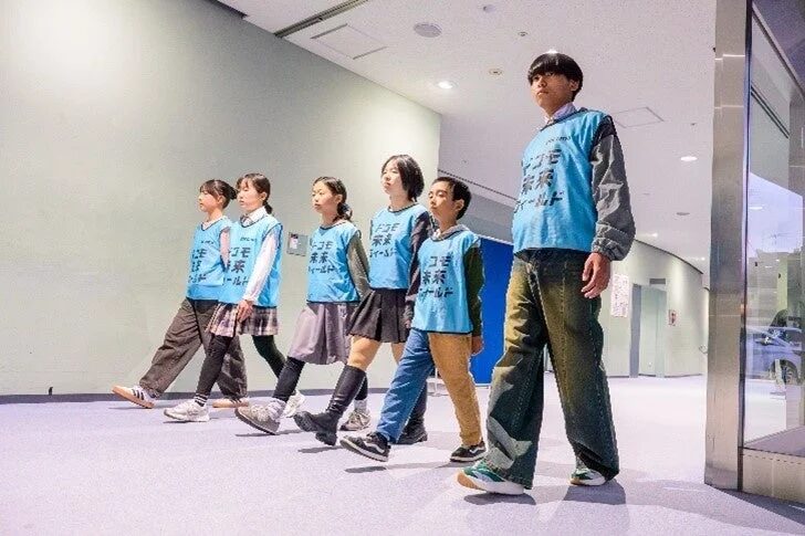 青いベストを着た子供たちが、屋内の広い空間で一列に並んで歩いている