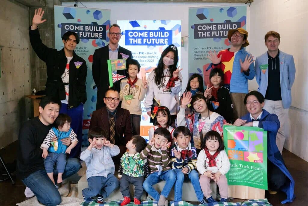 笑顔の大人と子供たちが集まる集合写真。背景には「COME BUILD THE FUTURE」のバナーと、大阪・関西万博のイギリス館に関連する「Park Track Picnic」のポスター