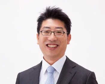 スーツと眼鏡を着用したアジア人男性がカメラに向かって笑顔を見せているポートレート写真です。