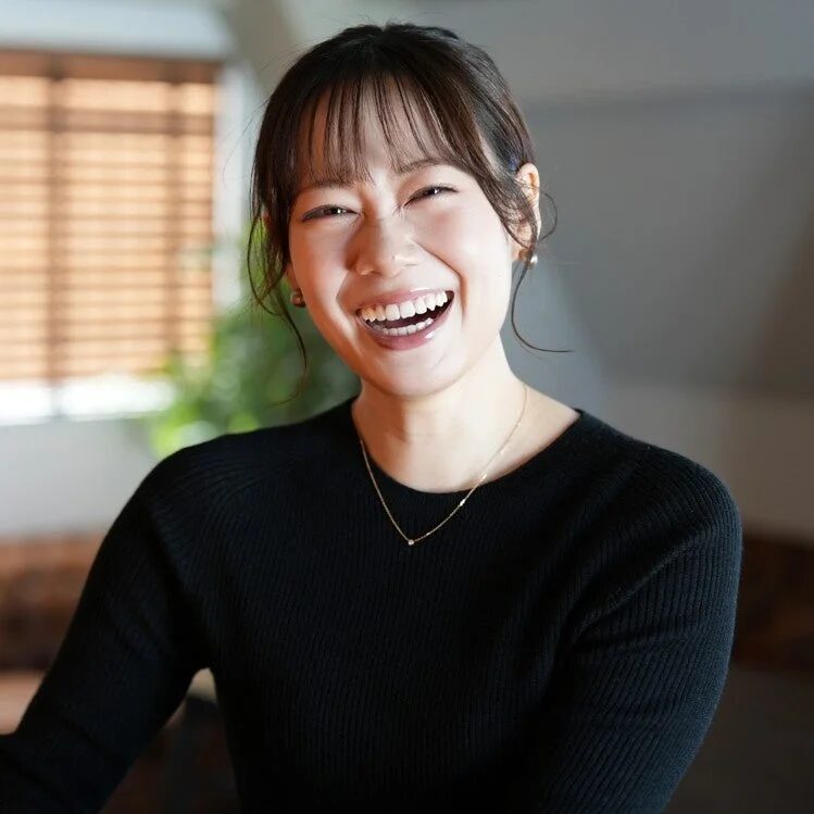 満面の笑みを浮かべた若い女性のポートレート。黒いトップスとシンプルなネックレスを身につけ、明るい自然光の中で幸せそうな表情を見せています。