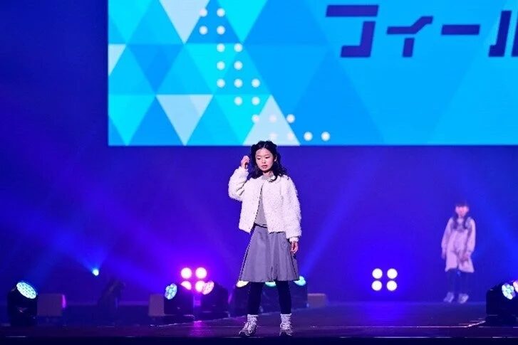 青い照明が当たるステージで、白いふわふわのジャケットと灰色のスカートを着用した少女がポーズをとっています