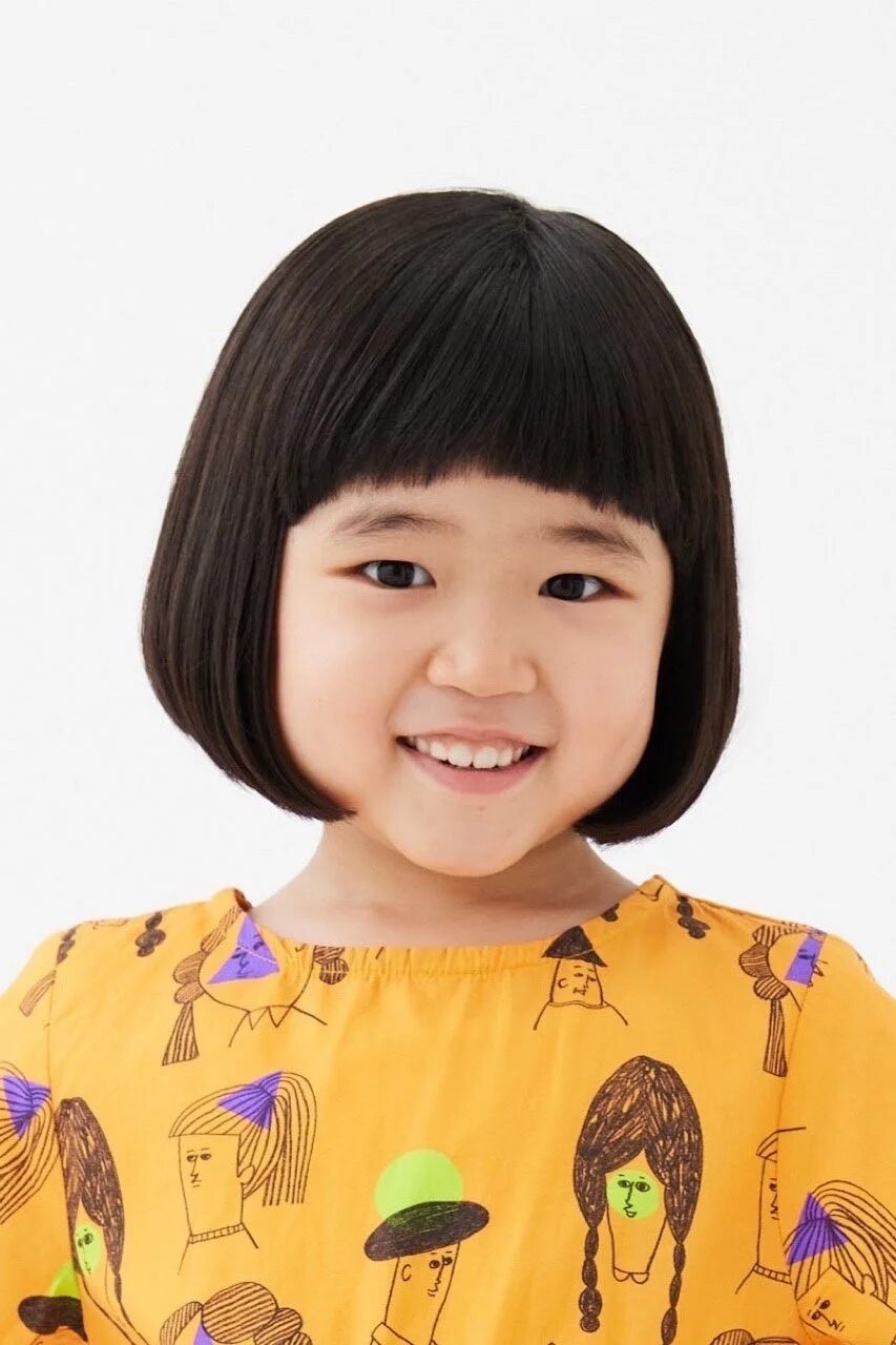 おかっぱ頭と前髪が特徴の、笑顔が可愛いアジア系の女の子のポートレートです。黄色の服にはユニークなイラストが描かれており、白い背景で撮影されています。