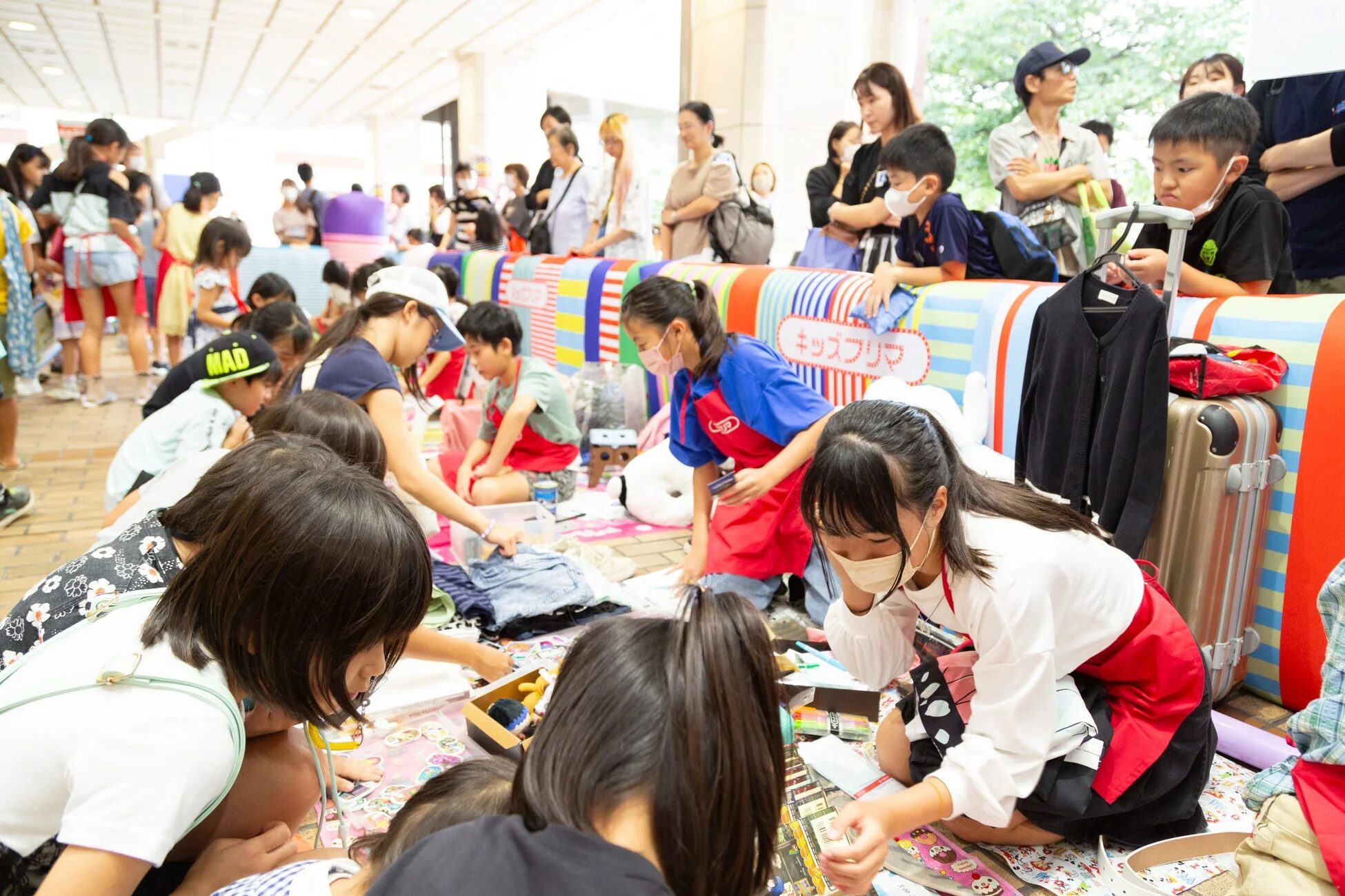 屋内のキッズフリマで、多くの子供たちがマスクとエプロンを着用し、活発に品物の売買や交流を楽しんでいる。