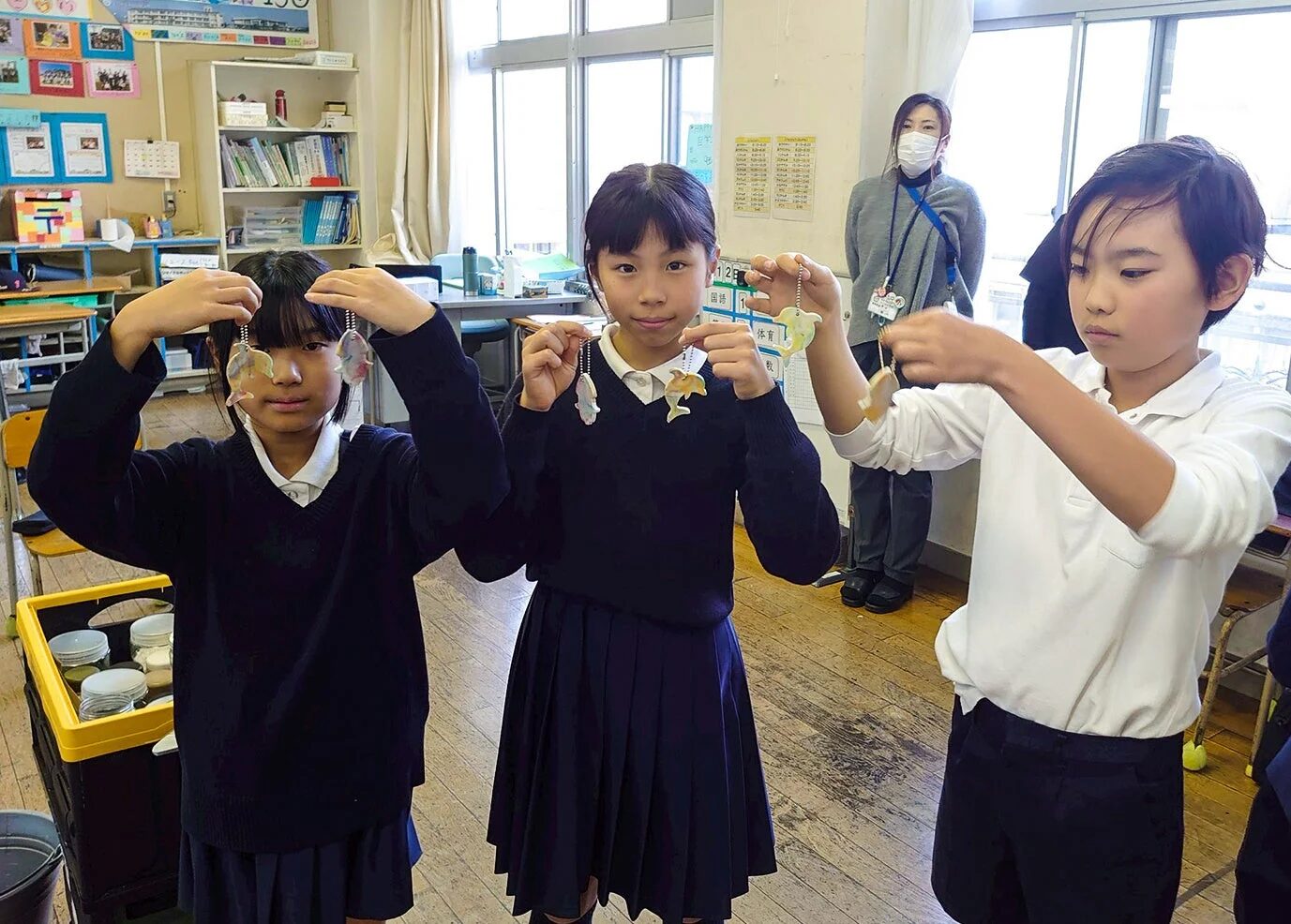 教室で3人の小学生が、自分たちで作ったと思われる魚やイルカの形をしたカラフルなキーホルダーを手にしている。