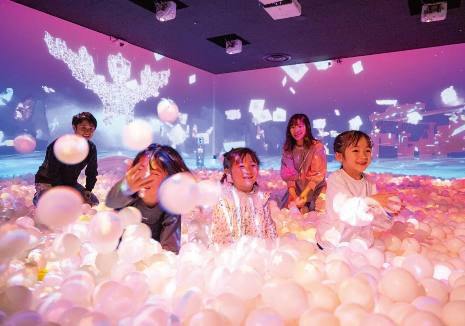 インタラクティブなデジタルアートが投影された空間で、子供たちが大人と一緒に光るボールプールで遊んでいます