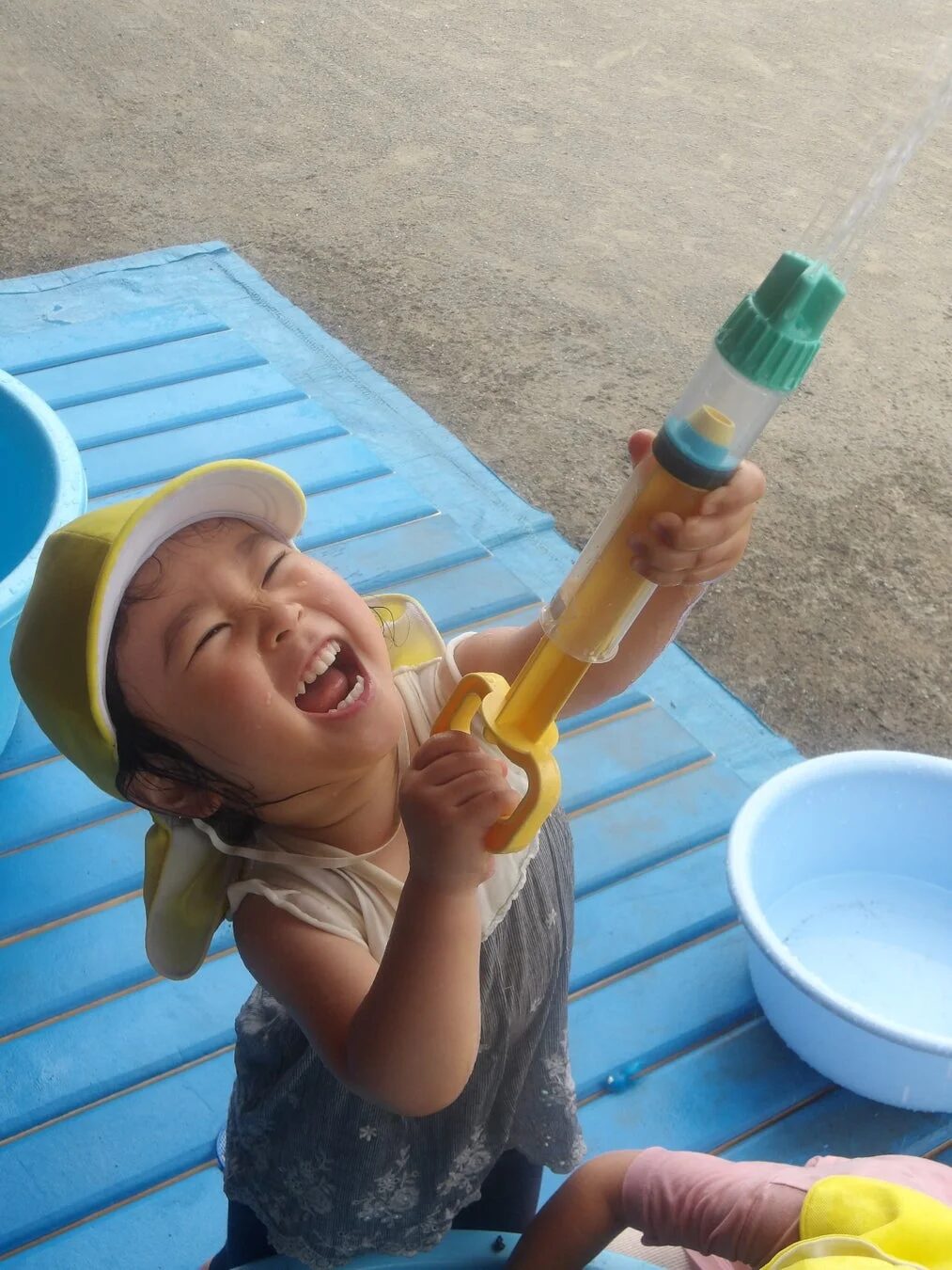 黄色い帽子をかぶった幼い子供が、水鉄砲で水を飛ばしながら満面の笑みを浮かべている。夏の屋外で水遊びを心から楽しんでいる様子が伝わる一枚。