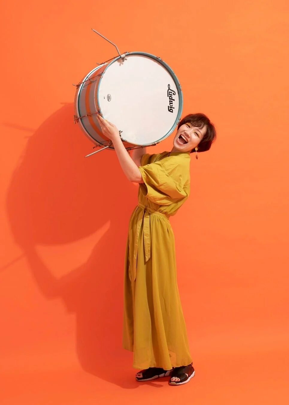 鮮やかなオレンジ色の背景の前で、黄色のワンピースを着た女性がドラムを抱え、満面の笑顔で楽しそうにしている
