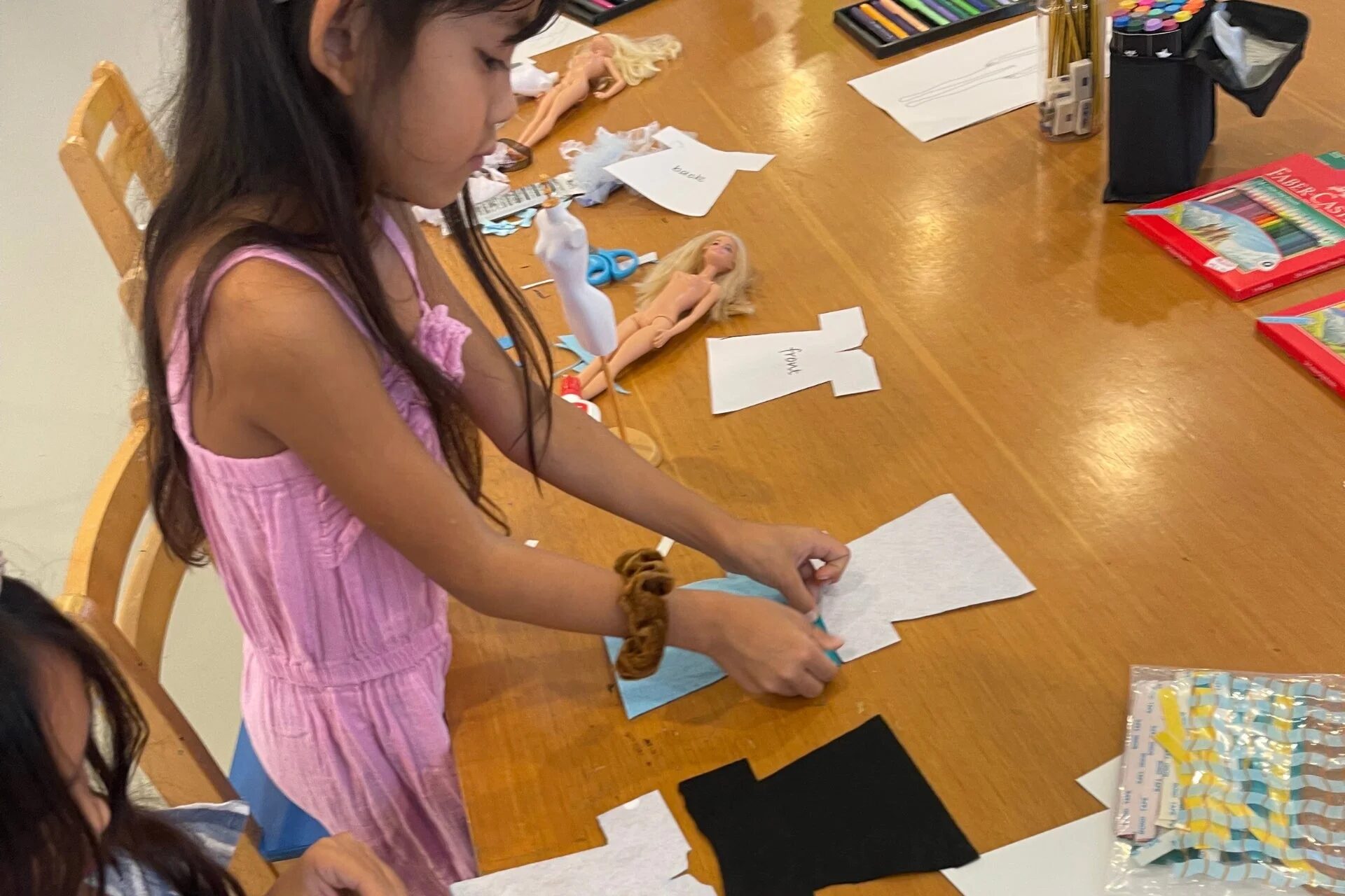 一人の少女が木製のテーブルでバービー人形の服を作る工作活動をしています。