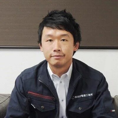 ダークブルーの作業服を着たアジア人男性が、カメラをまっすぐ見つめているポートレート写真です。背景はシンプルで、真面目な表情をしています。