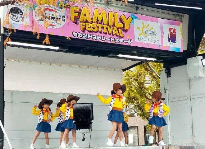 ファミリーフェスティバルのステージで、ウッディのようなカウボーイ衣装を着た子供たちが元気いっぱいにダンスを披露しています