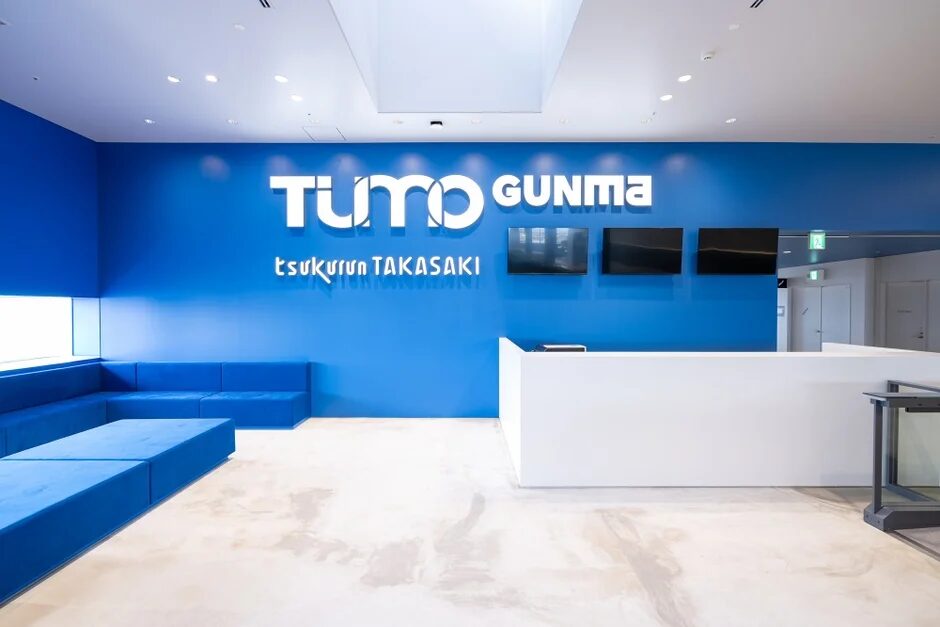 青い壁に「TUMO GUNMA」と「tsukurun TAKASAKI」のロゴが掲げられた、モダンで清潔感のあるオフィスの受付またはロビーエリア