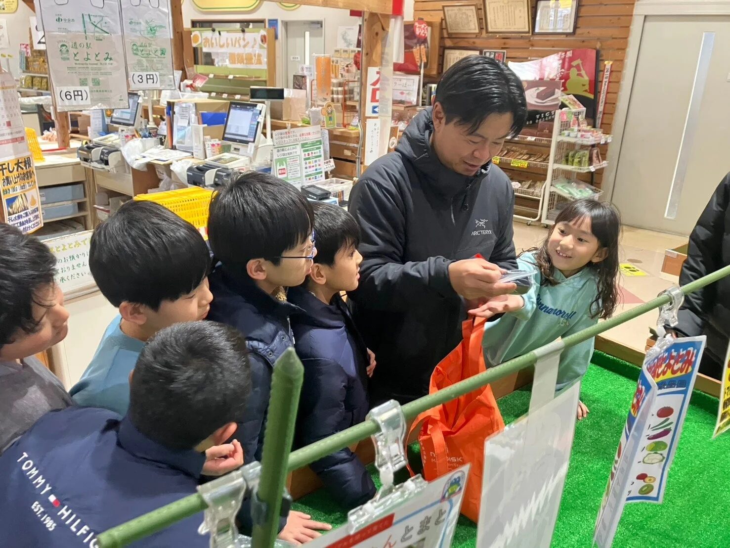 道の駅のような店内で、大人の男性が子供たちに何かを見せながら説明している