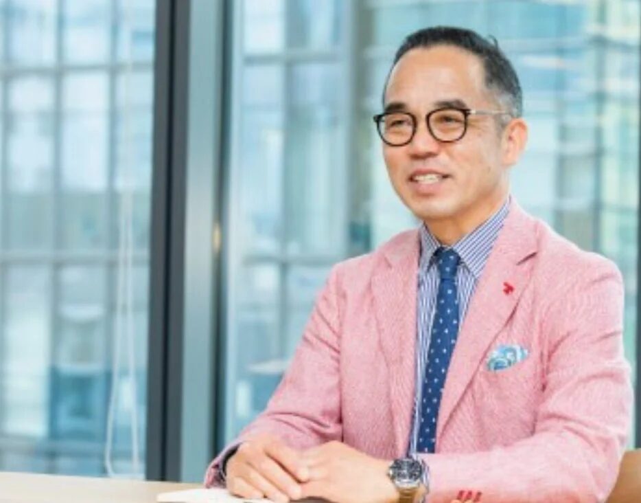 ピンクのジャケットとメガネをかけた男性が、オフィスのような場所で笑顔を見せています。