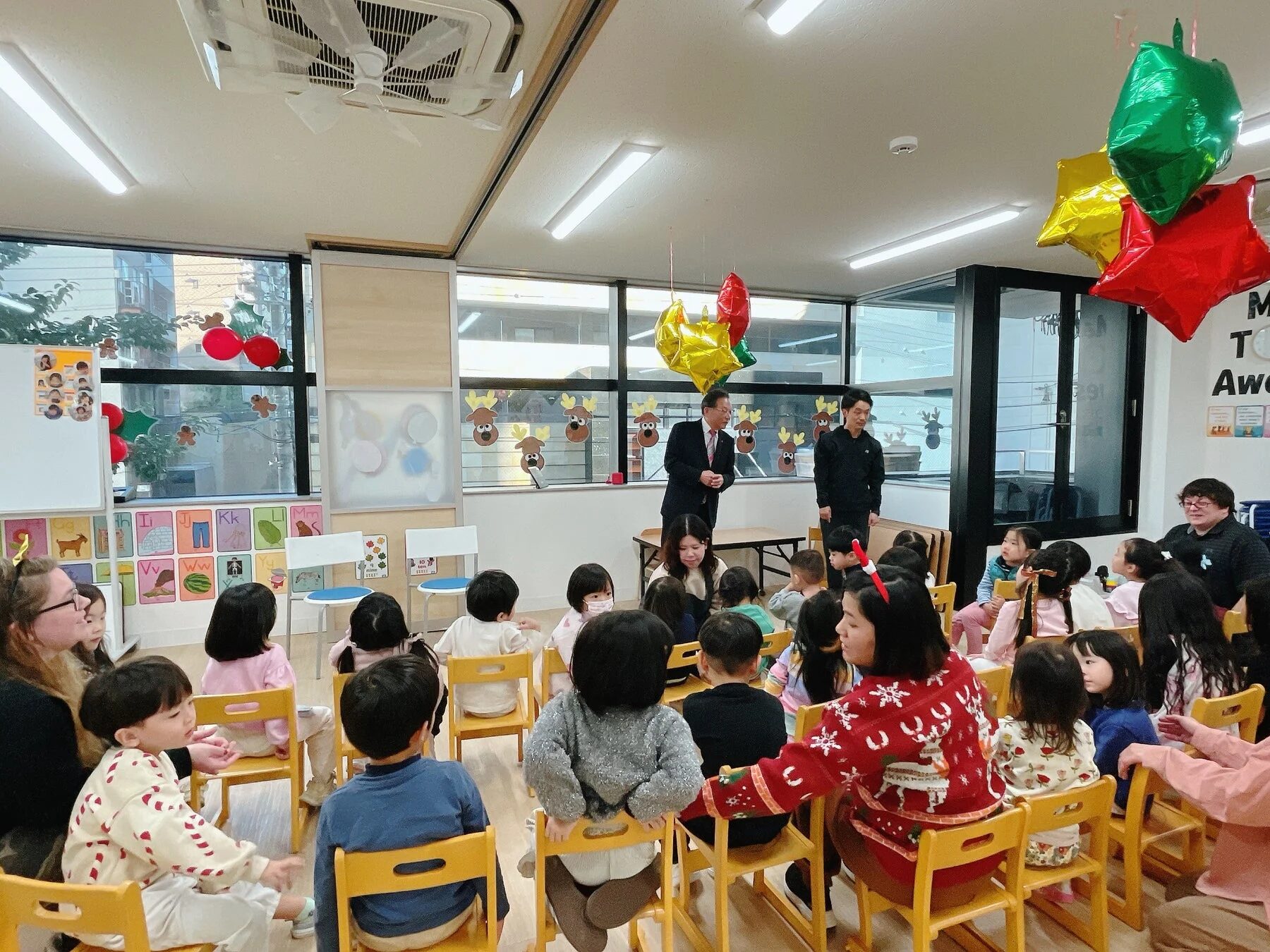 クリスマスをテーマにした飾り付けがされた教室で、多くの子どもたちが椅子に座って何かのイベントに参加している様子です。前方に立つ2人の男性が話をしており、数人の大人が子どもたちを見守っています。