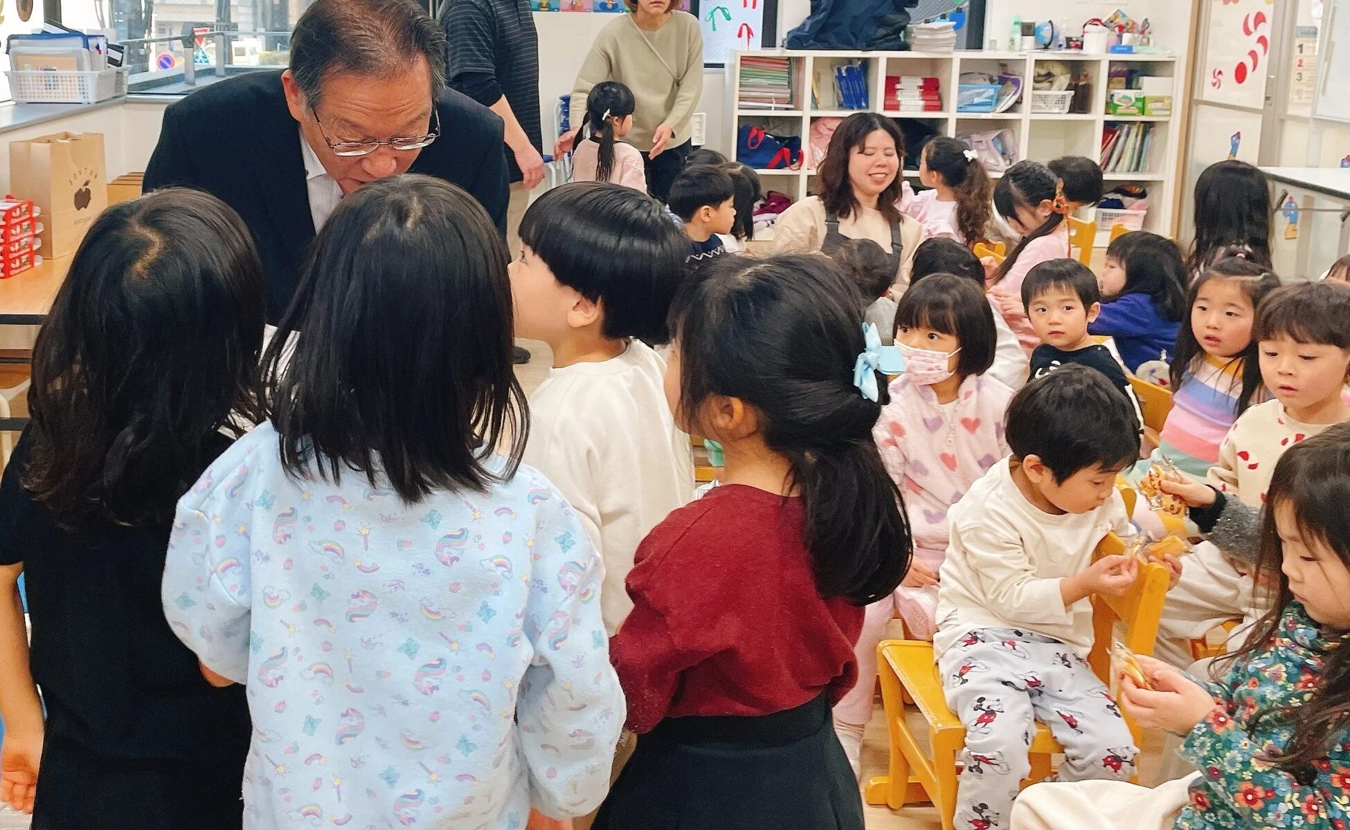 幼稚園または保育園で、スーツ姿の男性が子供たちと交流している様子です。多くの園児が集まり、男性からの配布物らしきものを受け取ったり、楽しそうに見つめたりしています。和やかな雰囲気のイベントのようです。