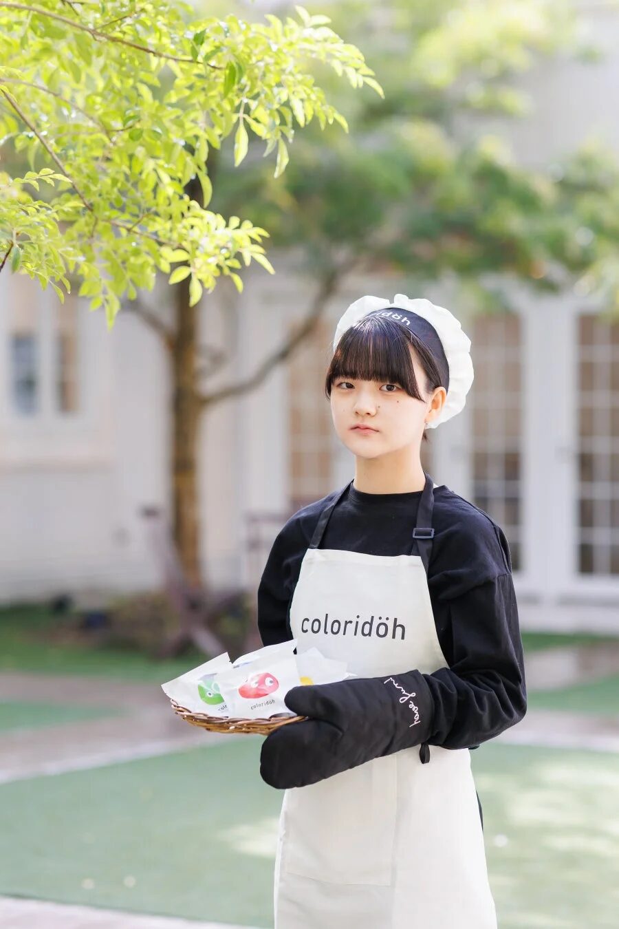 エプロンと帽子を着用した若い女性が、屋外の緑豊かな場所でバスケットに入ったカラフルなパッケージ商品を手に持っている