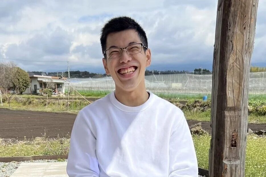 眼鏡をかけた若い男性が白いスウェットシャツを着て、屋外で満面の笑みを浮かべているポートレート写真です。背景には農村の家屋や畑、ビニールハウスが見え、曇り空の下でリラックスした雰囲気です。