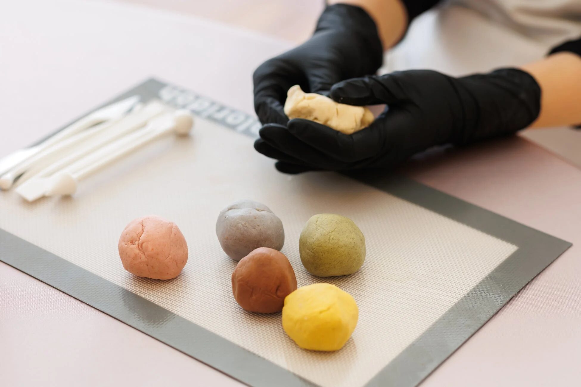 黒い手袋をはめた手が生地をこねており、シリコンマットの上には様々な色の丸い生地が並べられている
